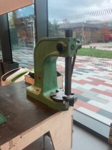 A painted pale green cast iron arbor press sat on the edge of a metal-topped workbench. In the background there is a window looking out onto a paved area and green grass.